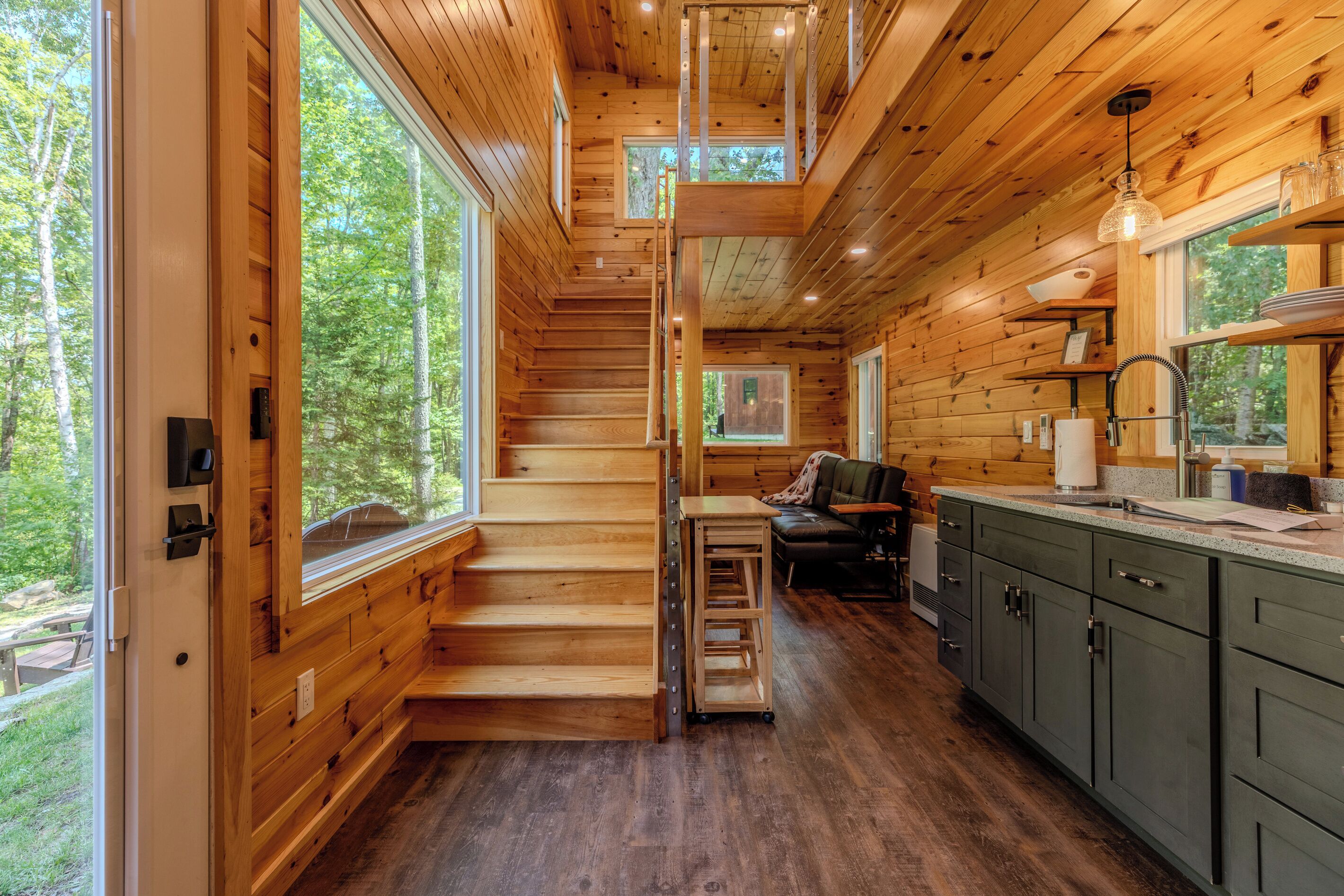 L’intérieur d’une cabane en bois est aménagé avec un escalier menant à une mezzanine, un coin cuisine et de grandes fenêtres qui offrent une vue sur la forêt environnante.
