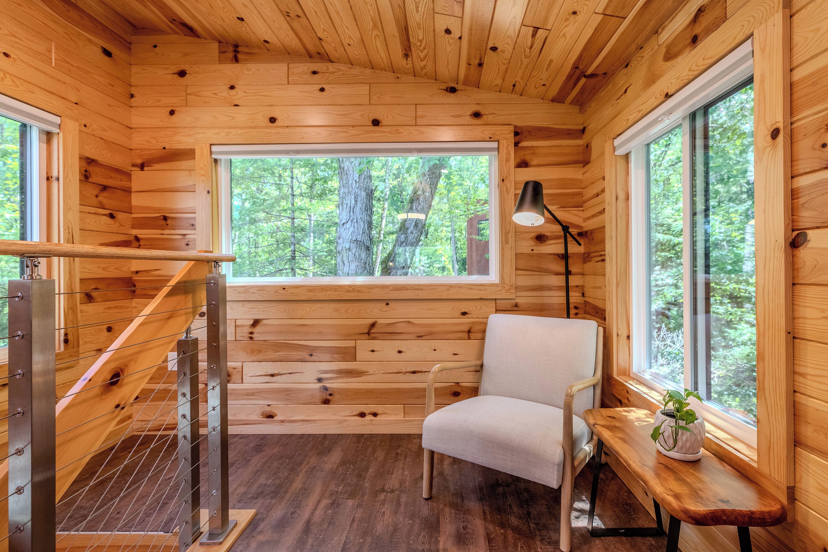 Ein Interieur einer Holzhütte mit einer Holztreppe, einem einzelnen Sessel, einem kleinen Tisch mit Pflanze und einem großen Fenster mit Blick auf Bäume.