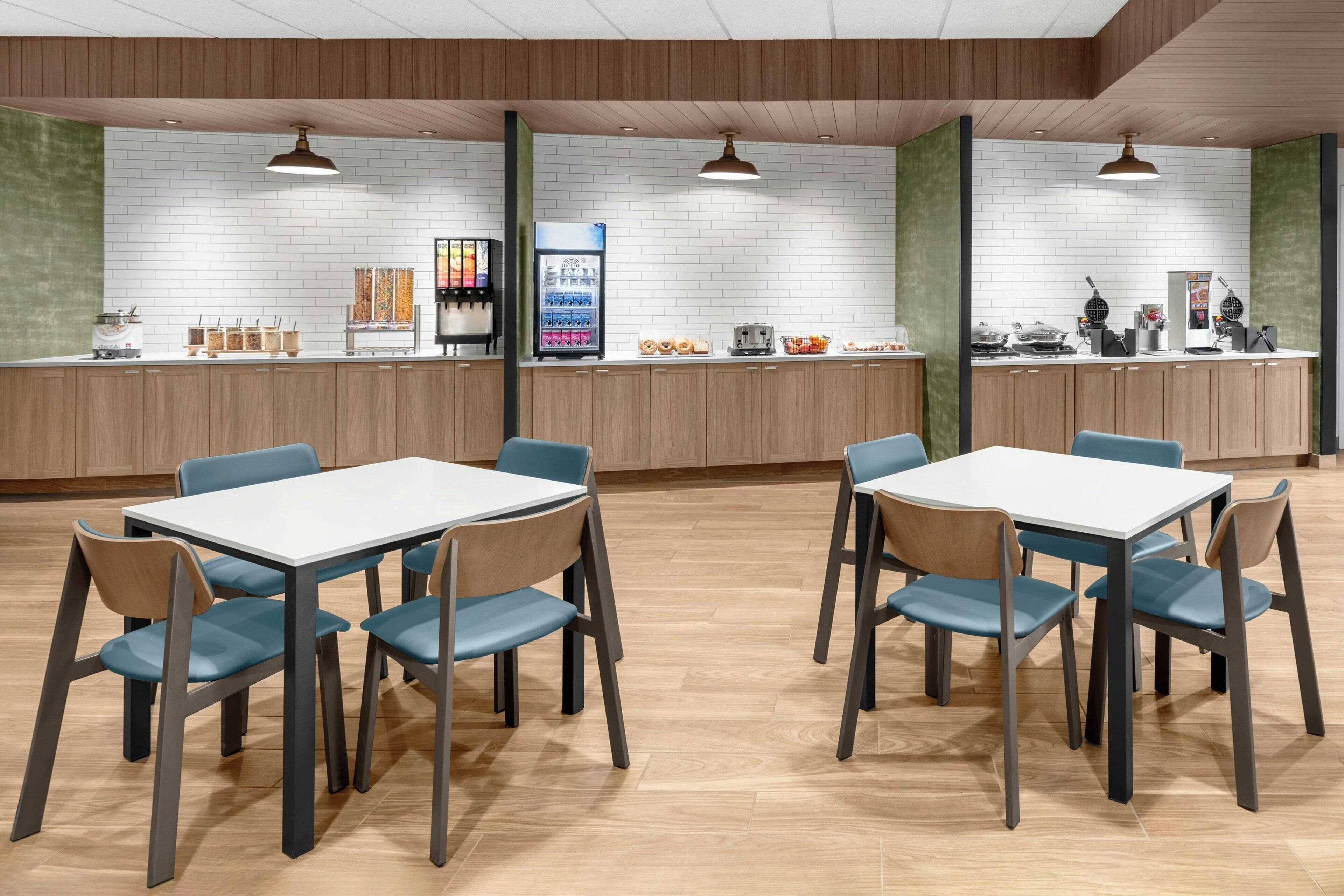 A modern dining area with two tables and chairs, a counter with a beverage dispenser, and a kitchen area in the background.