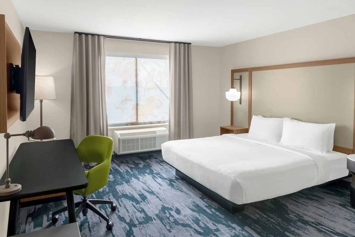 Habitación de hotel con cama, escritorio, silla, lámpara y ventana con cortinas.