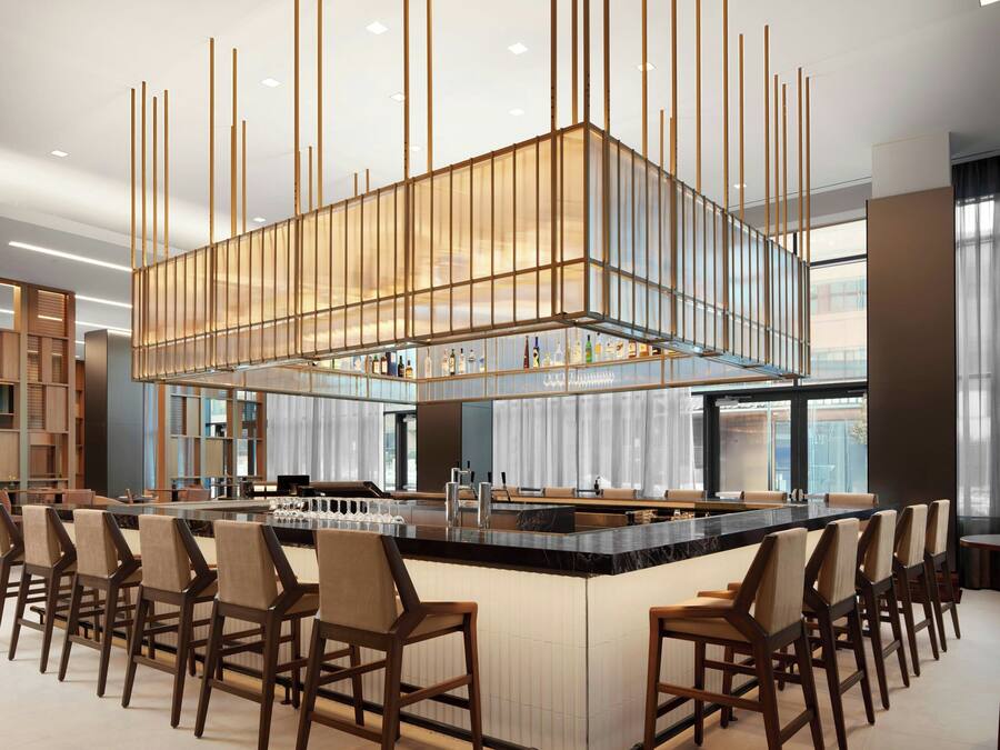 Un espace bar moderne doté d’un îlot central, de tabourets de bar et d’un plafond haut orné de luminaires verticaux.