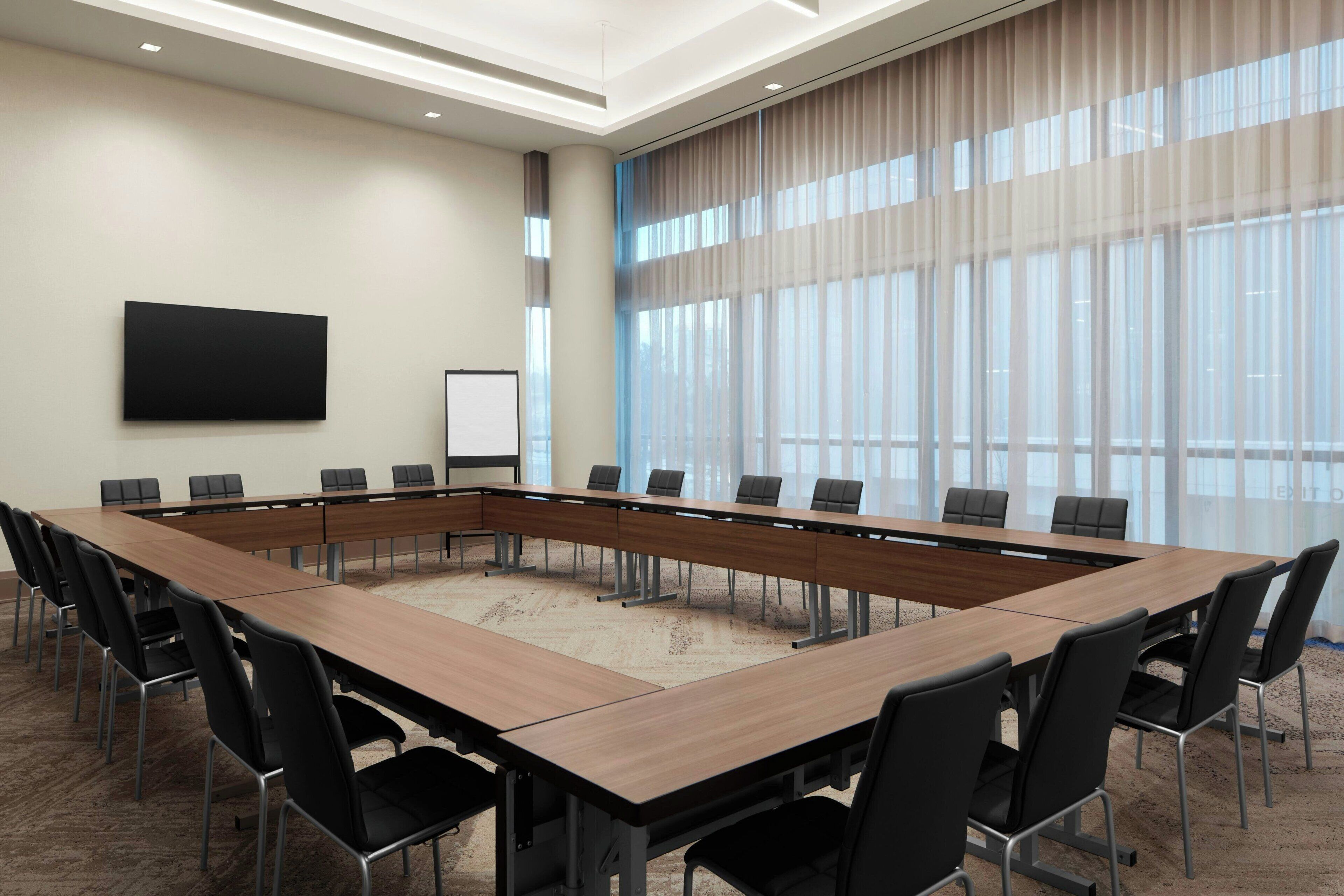 Un salón de conferencias con una larga mesa, sillas negras, un televisor de pantalla plana y amplias ventanas con cortinas translúcidas.