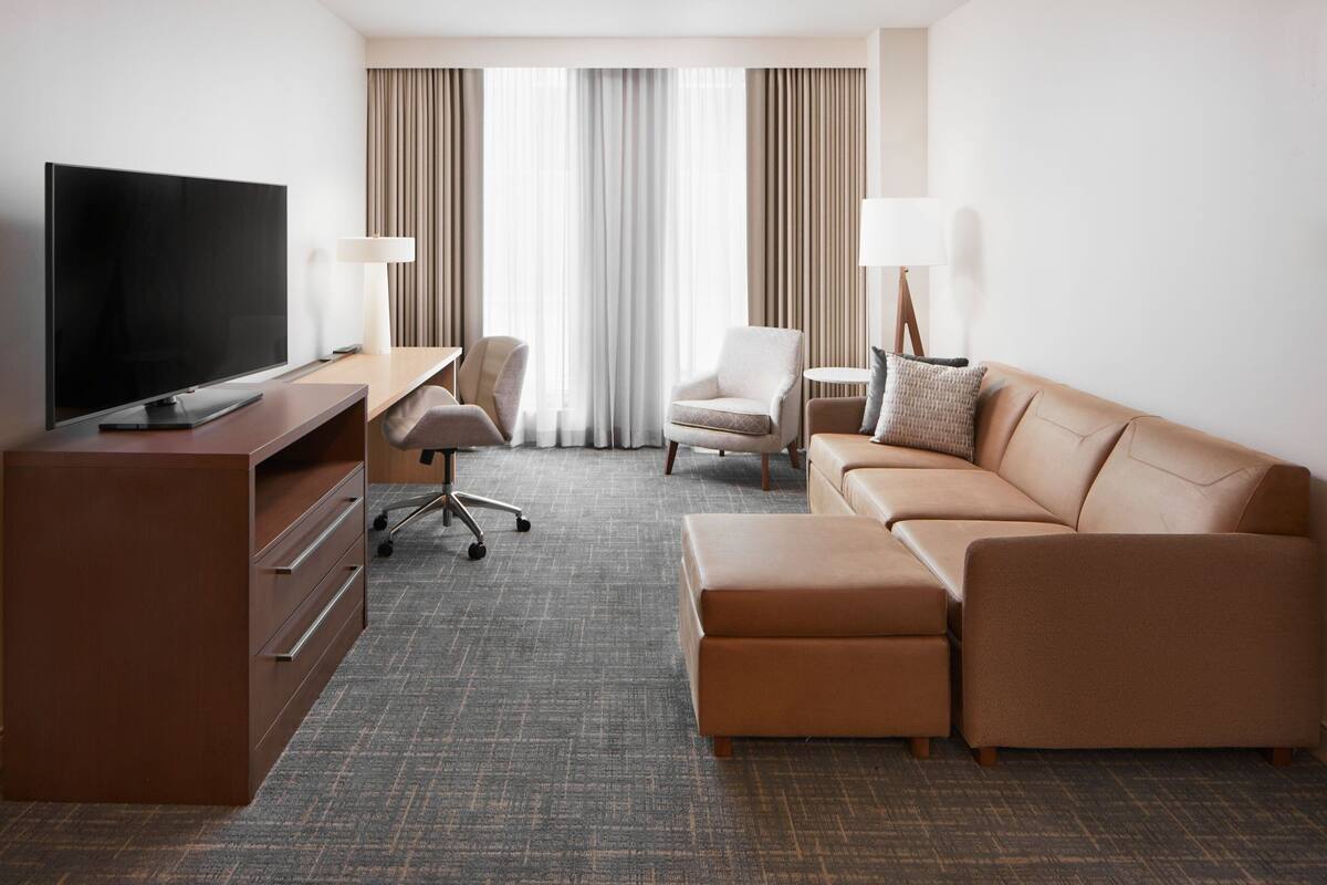 A modern hotel room with a flat-screen TV, a wooden desk, a leather sofa, a small armchair, and a large window with curtains.