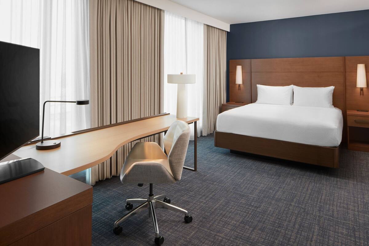 A hotel room with a bed, desk, chair, lamp, and TV.