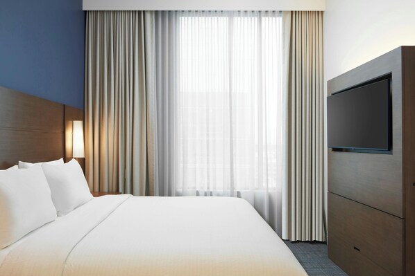 Habitación de hotel con una cama grande, televisor montado en la pared y una ventana con cortinas de gasa.