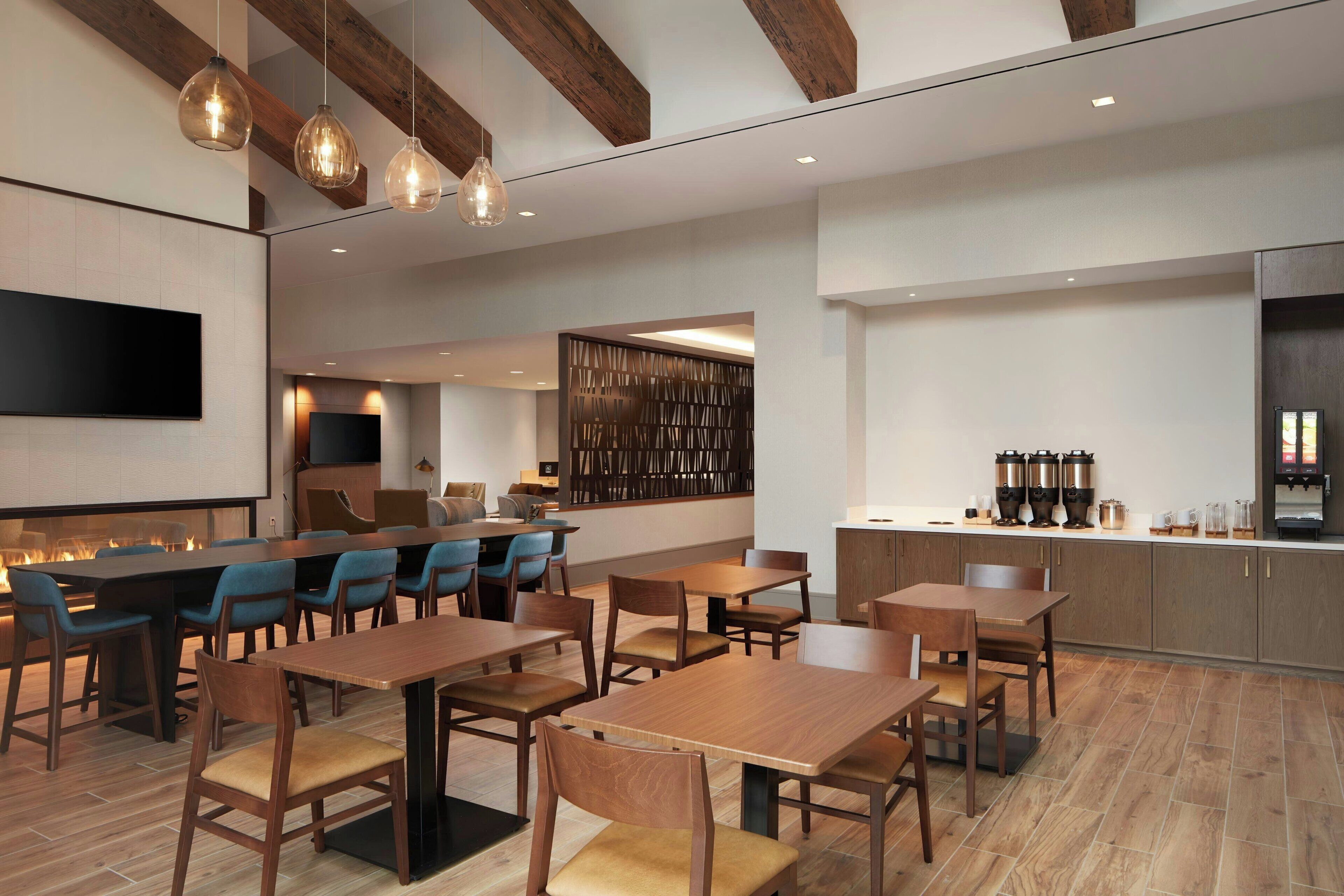 A modern restaurant with wooden tables and chairs, a bar area, and a kitchen visible in the background.