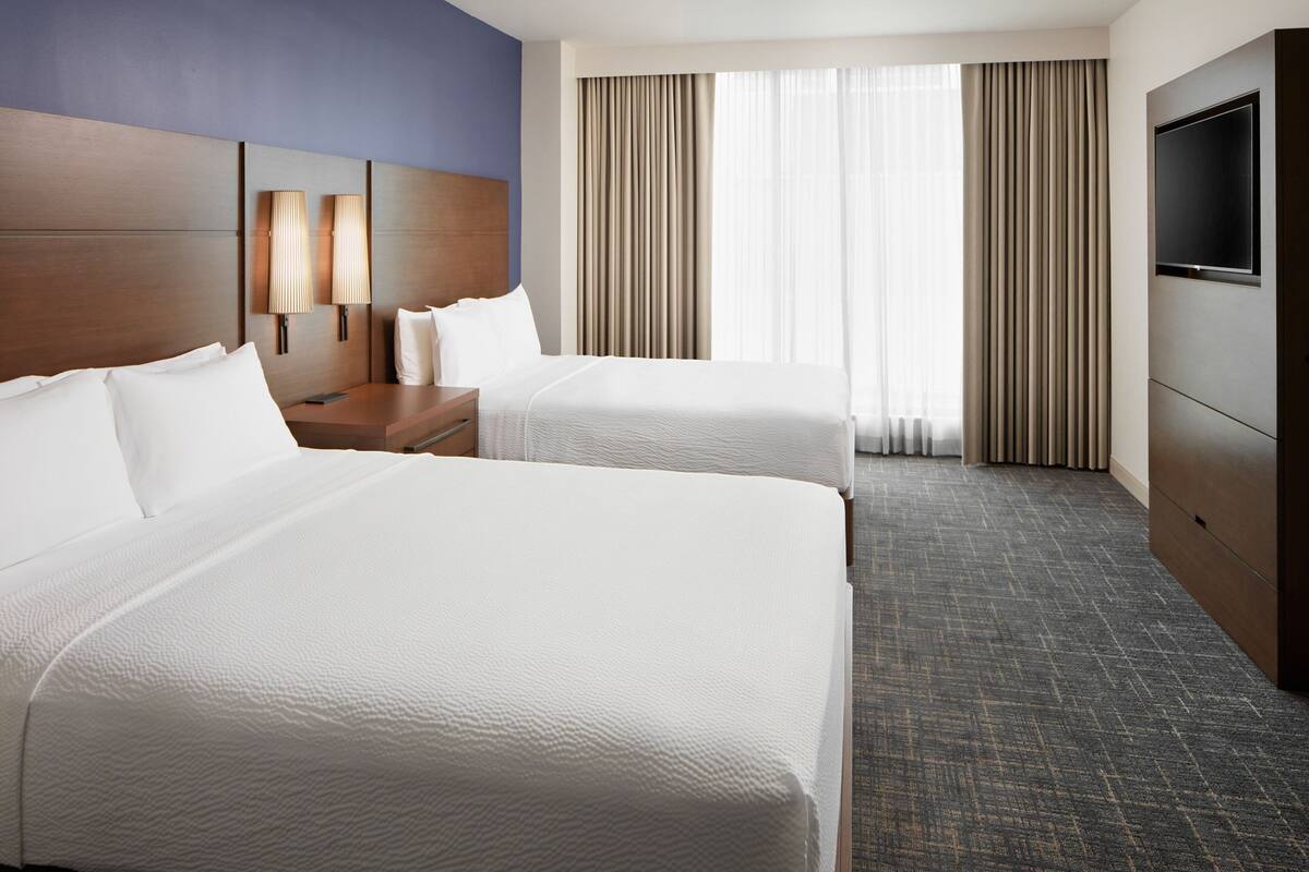 A hotel room with two beds, a wooden headboard, a flat-screen TV, and a large window with curtains.