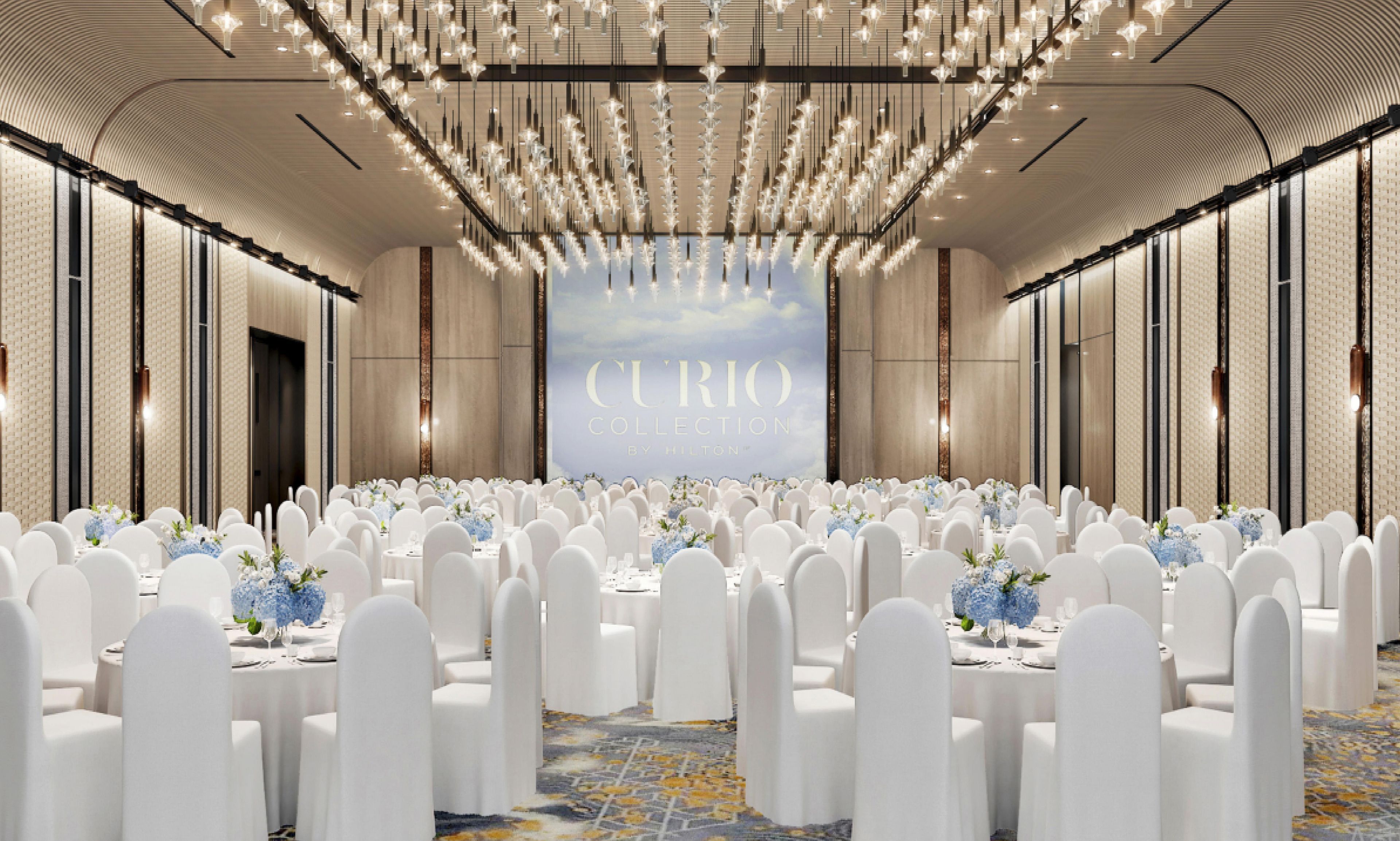Un amplio y bien iluminado salón de banquetes con filas de mesas redondas dispuestas para un evento formal, con un fondo que muestra el texto 'CURIO COLLECTION BY HILTON'.