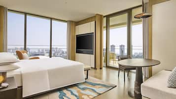 A hotel room with a large bed, a TV, a dining table, and a balcony with a city view.