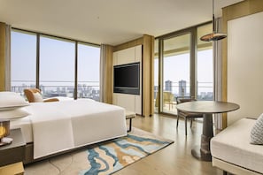 Habitación de hotel con una cama grande, televisor, mesa de comedor y balcón con vista a la ciudad.