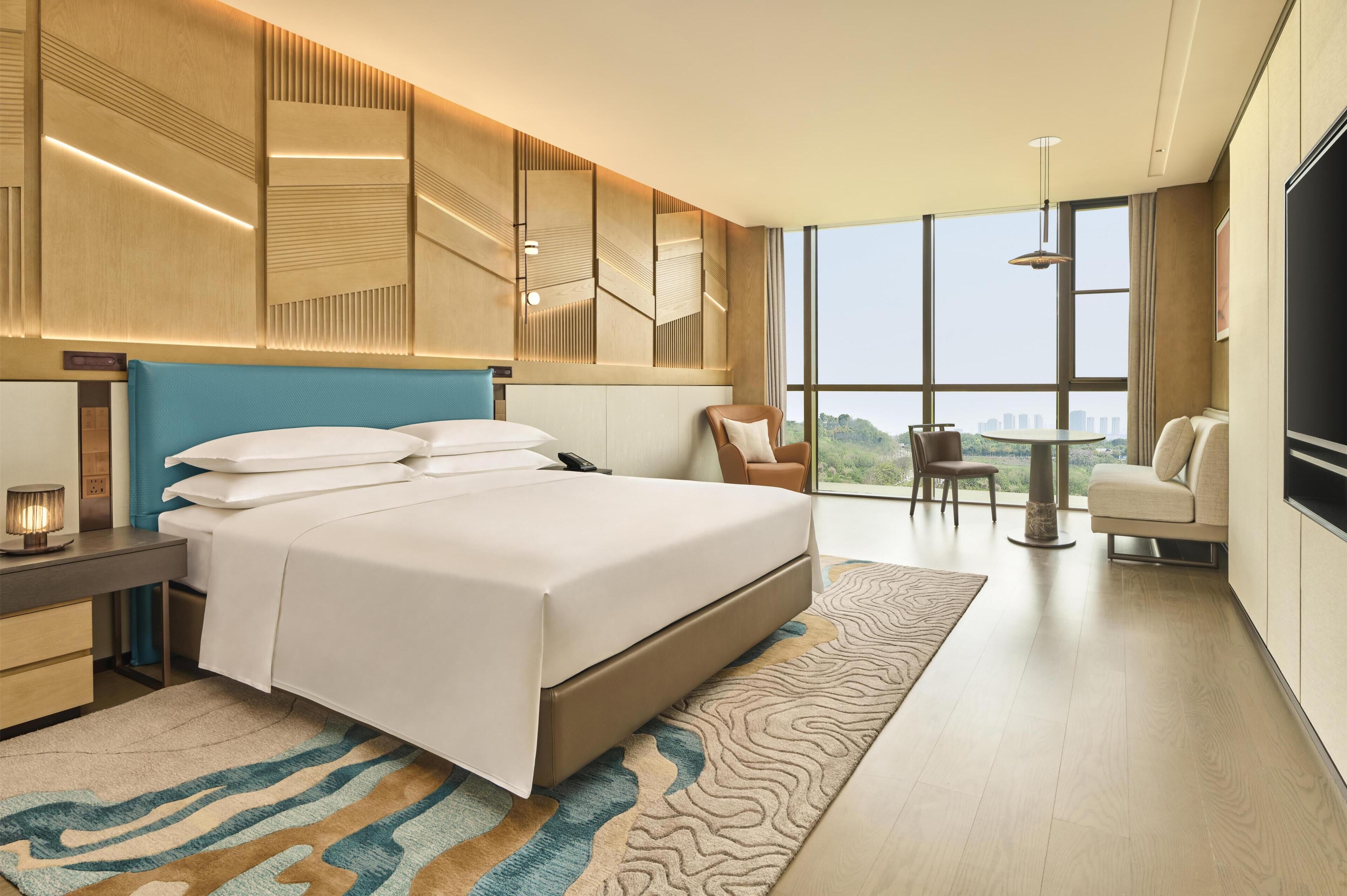 Quarto de hotel moderno com uma cama grande, uma escrivaninha, uma cadeira, uma televisão e vista para a cidade.