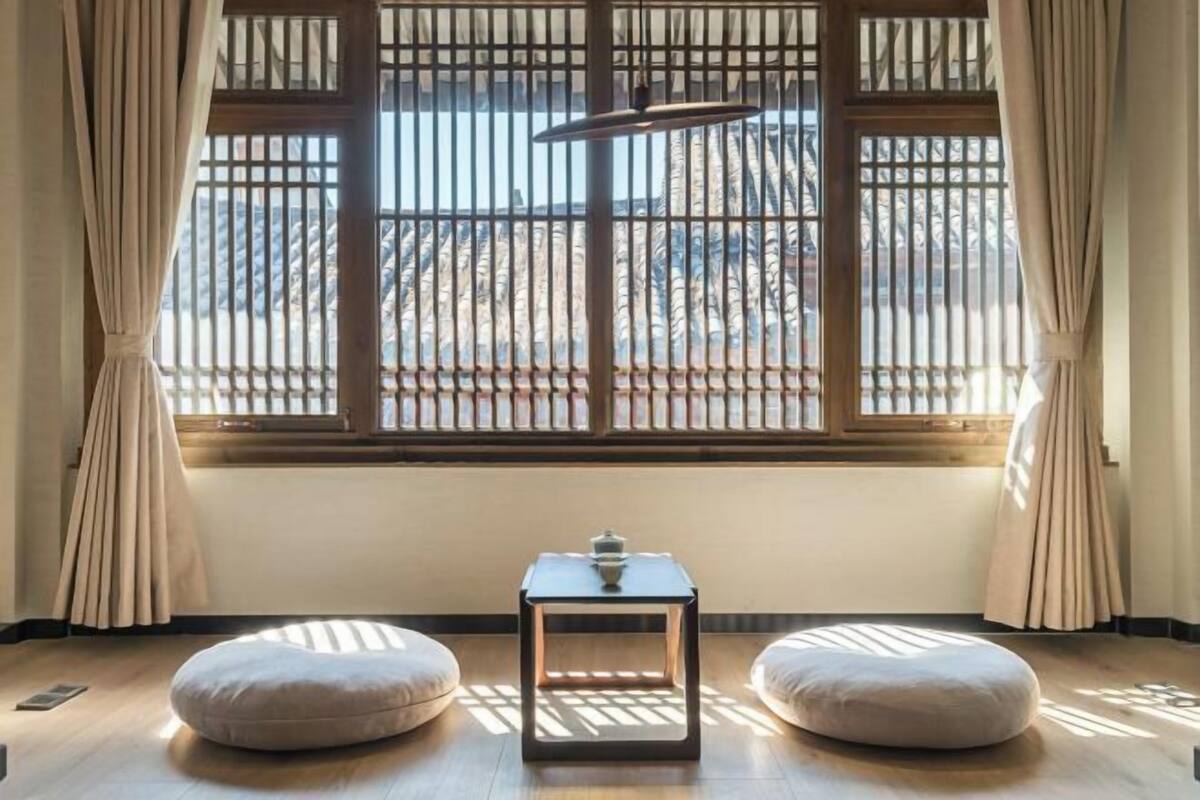 Una habitación con piso de madera, una mesita con lámpara, dos sillones de peluche y un ventanal con persianas verticales.