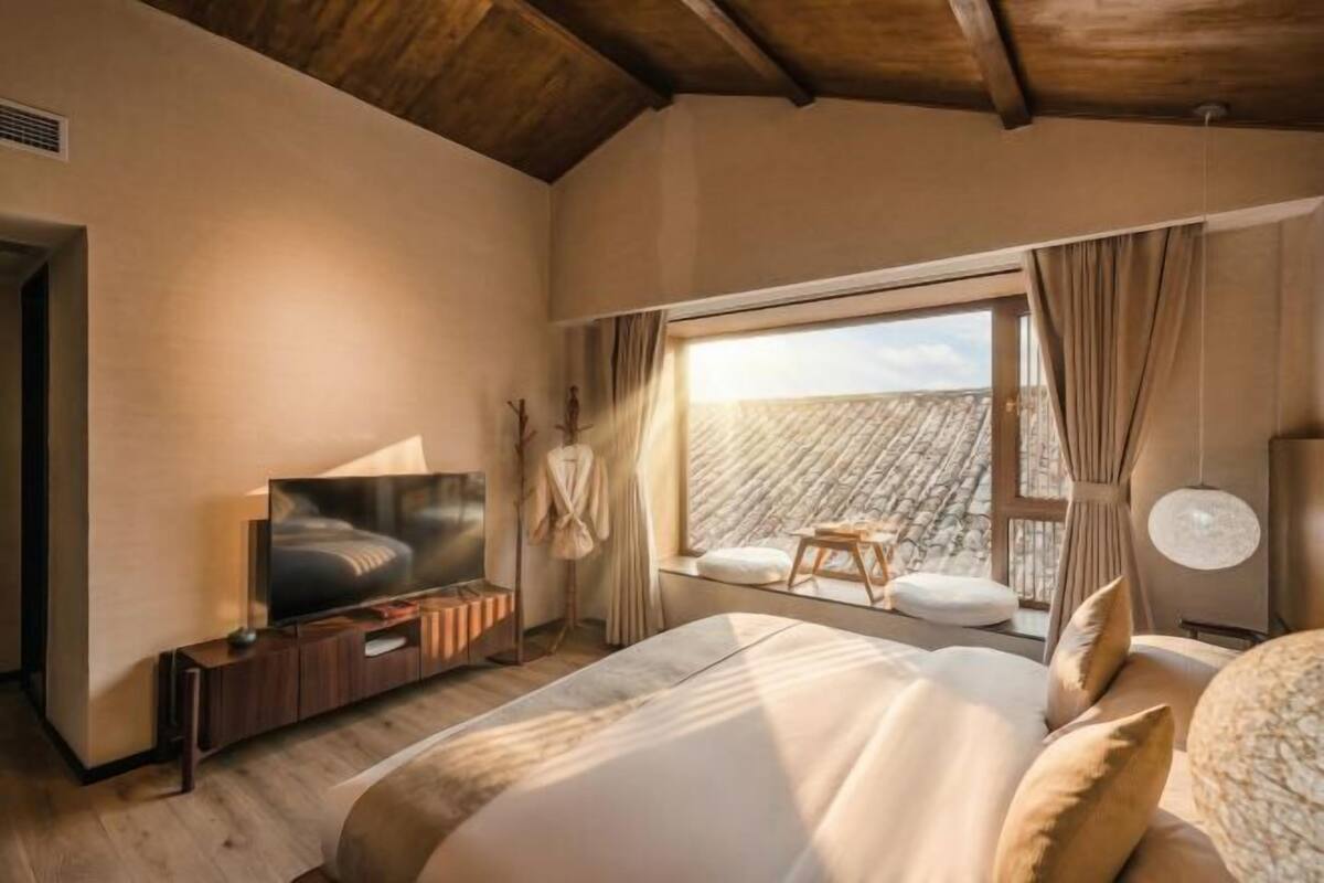 Un dormitorio con una cama grande, televisión, ventana con cortinas y vista al exterior.
