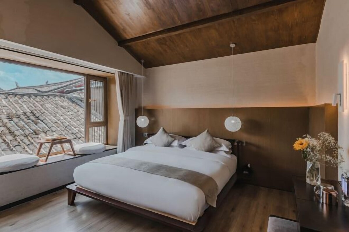 A bedroom with a large bed, wooden ceiling, and a view of a traditional roof through a window.