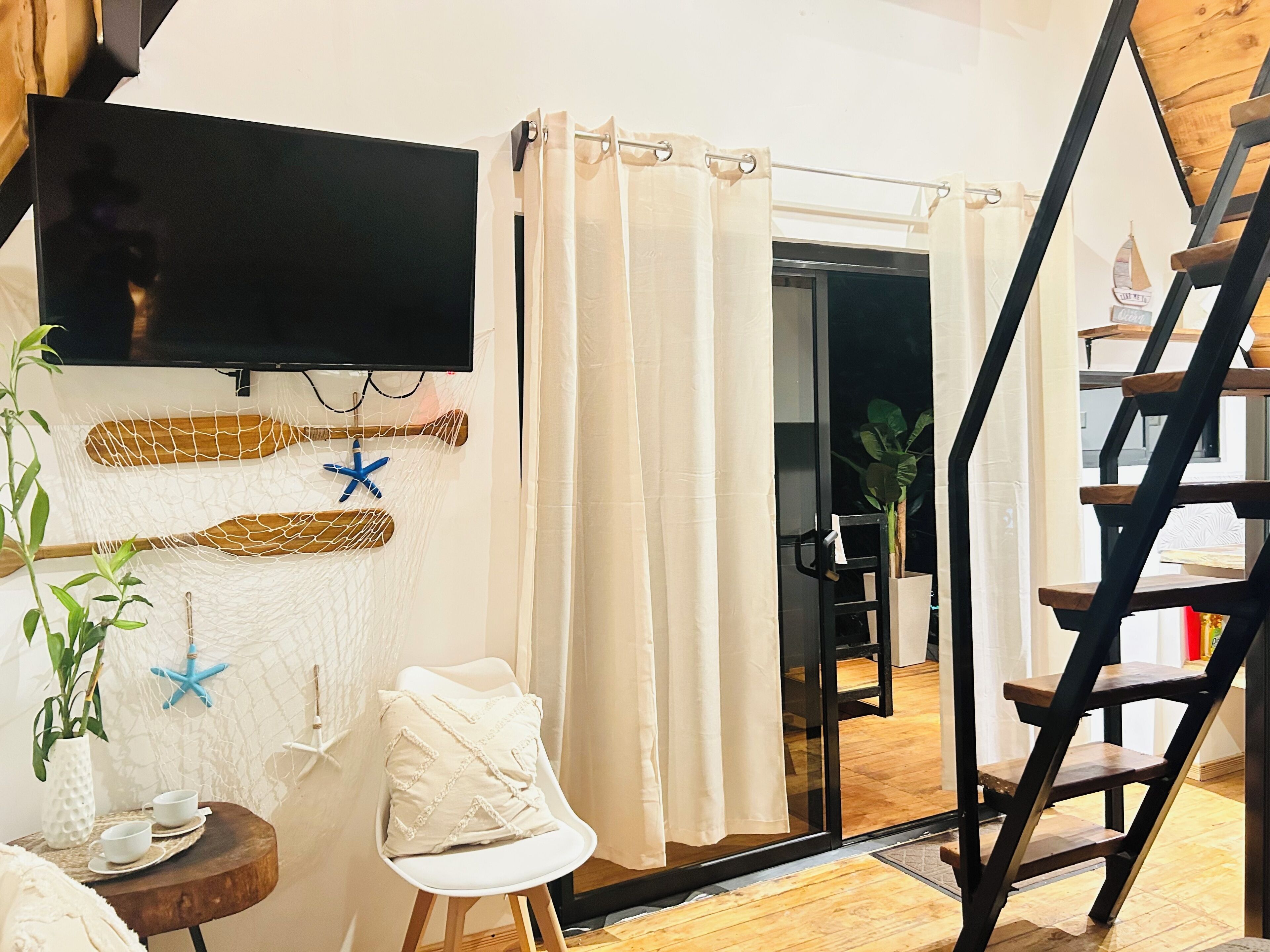 A room with a TV mounted on the wall, a white chair, a small table with cups, a wooden ladder, and a door leading to another room.