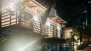 Une structure en bois à plusieurs niveaux, dotée de balcons, éclairée par un système d’éclairage artificiel et entourée de végétation.