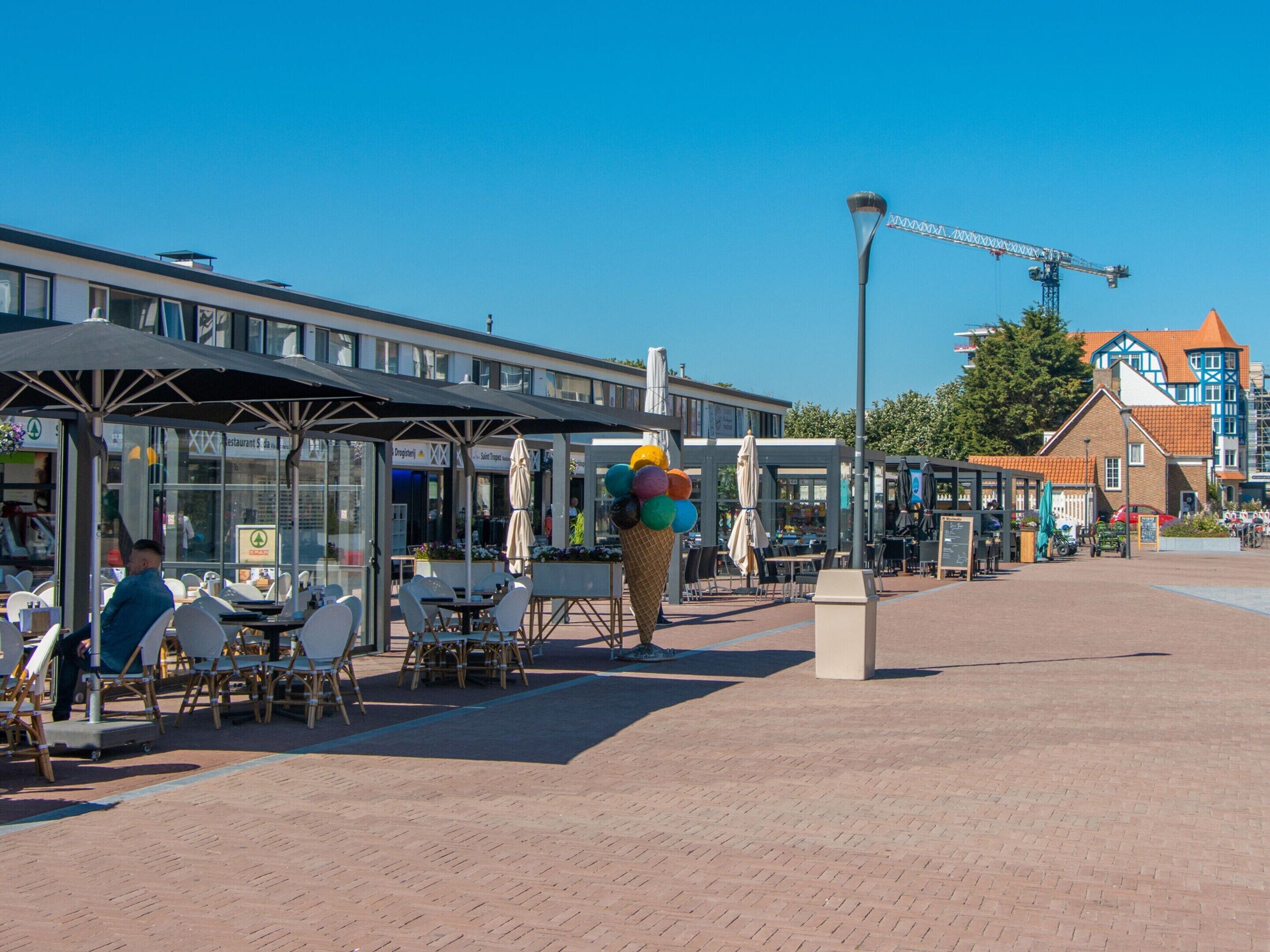 A paved outdoor area with tables and chairs under umbrellas, a large ice cream cone, and a street with buildings in the background.