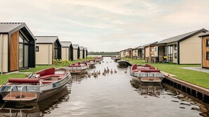 A canal with boats docked on either side, surrounded by modern buildings with large windows and a grassy area in the foreground. - Fenced Garden Family Chalet (Giethoorn)