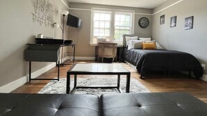Quarto compacto com cama, escrivaninha, cadeira, televisão e janela.