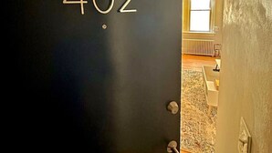 Una puerta negra con el número 402 que da a una habitación con ventana, radiador y una silla blanca.