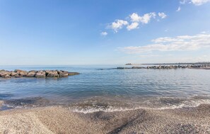 Una playa de canto rodado con aguas cristalinas, un rompeolas rocoso y una costa lejana con edificios.