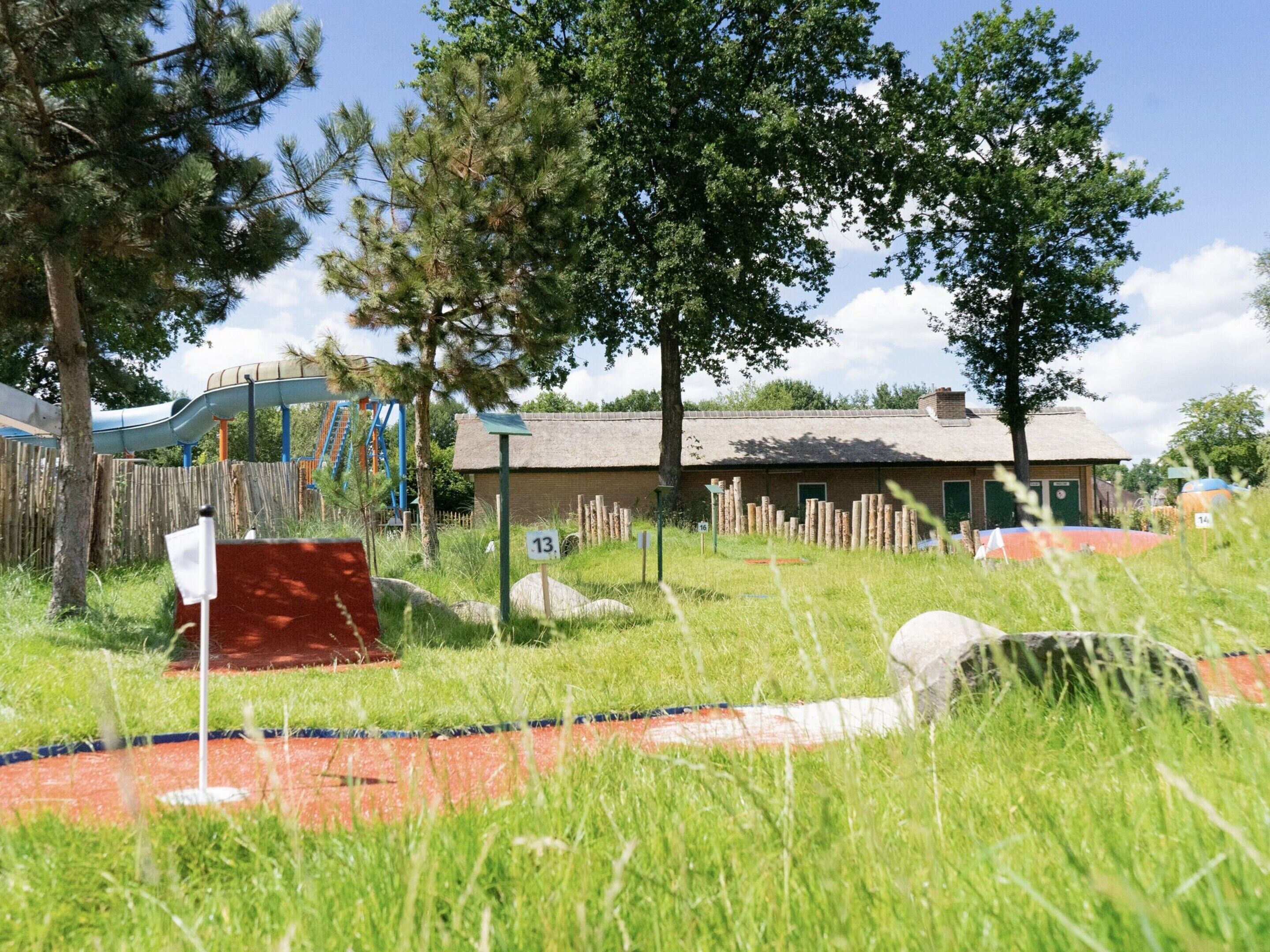 A rustic building with a thatched roof, surrounded by tall trees and a playground with a slide.