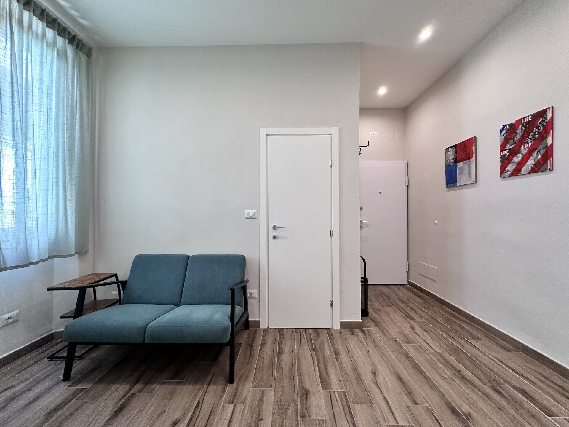 A modern living room with a blue sofa, wooden flooring, a white door, and two artworks on the wall.