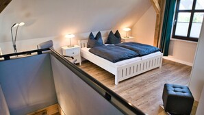 A bedroom with a sloped ceiling, a white bed with dark bedding, two bedside lamps, and a small black ottoman. - Baltic Sea Barn \"otto\" (Luessow)