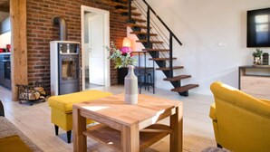A modern living room with a wooden table, yellow ottoman, and a vase with flowers. A staircase with a black railing is visible in the background. - Baltic Sea Barn \"otto\" (Luessow)