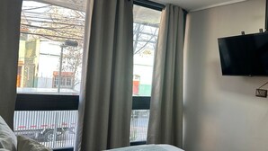 Quarto de hotel com cama, cortinas, televisão fixada na parede e janela com vista para a rua.