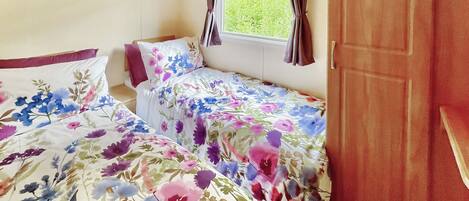 A small bedroom with two beds, wooden cabinets, a window with curtains, and a view of greenery outside.