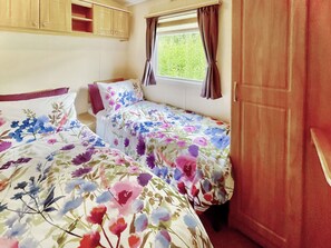 A small bedroom with two beds, wooden cabinets, a window with curtains, and a view of greenery outside. - Shirley's Haven - UK46631 (Driffield)