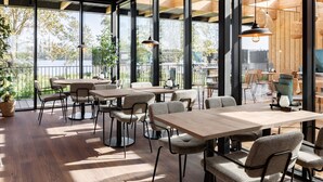 A modern dining area with wooden tables and chairs, large windows offering views of trees and a body of water. - Luxurious Lodge in a Holiday Park (Zwartewaal)
