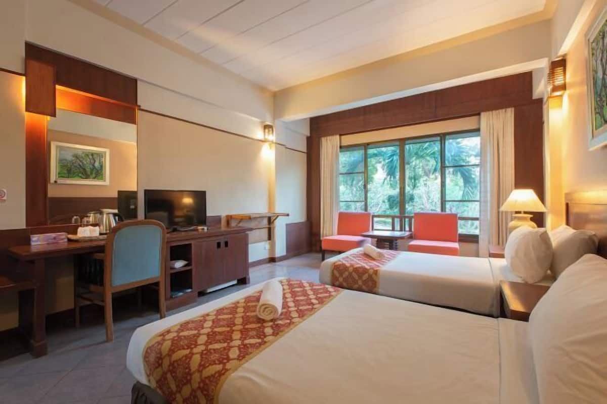 A hotel room with two beds, a desk, a television, and a window with a view of greenery.