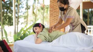 A person receiving a massage on a bed outdoors with a brick wall and greenery in the background.