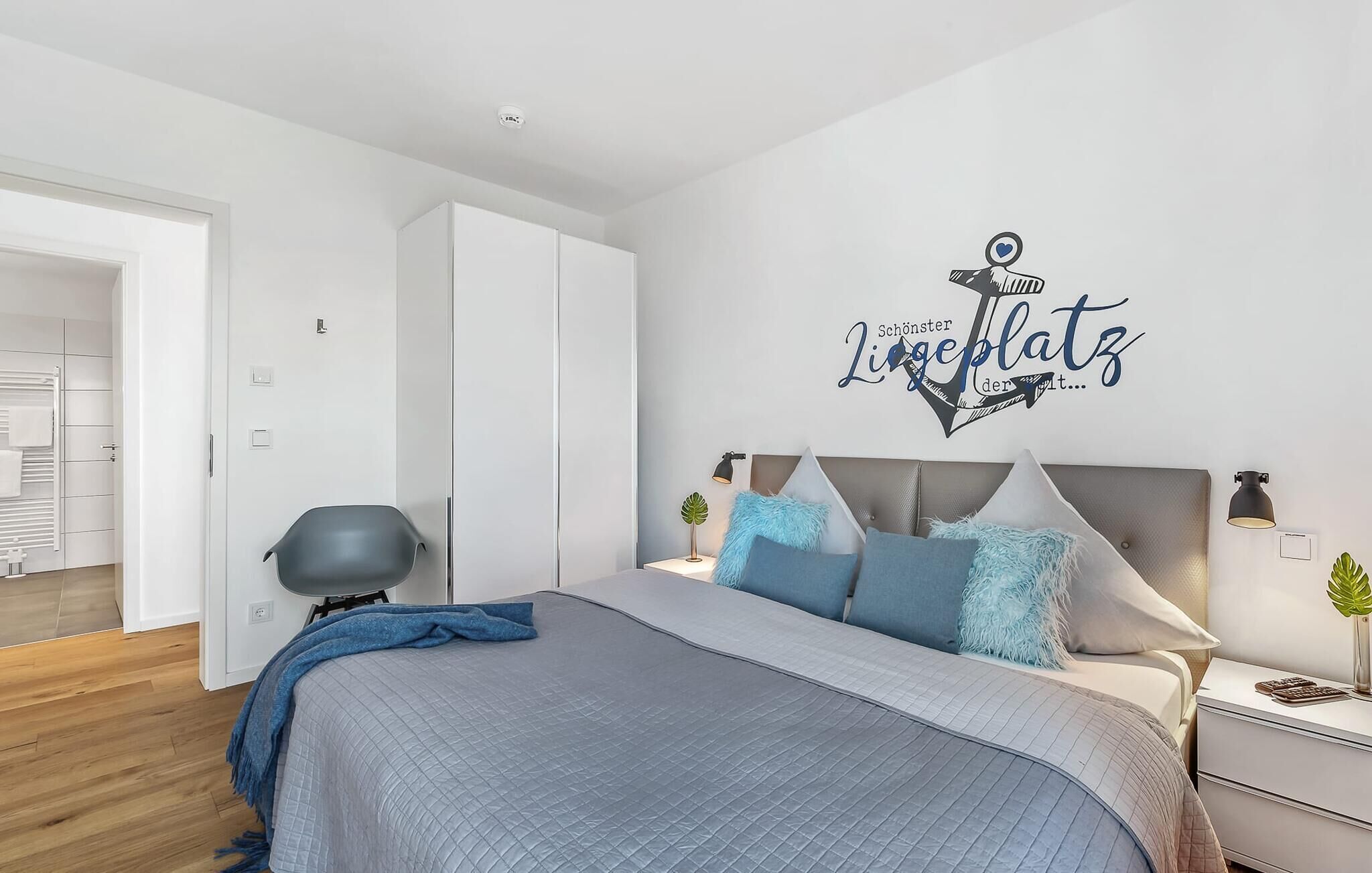 A modern bedroom with a bed, bedside tables, a nightstand, a wall-mounted light, and a decorative anchor wall art.
