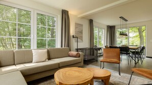 Una sala moderna con un sofá, dos mesitas de madera, un comedor con mesa y sillas, y ventanas amplias con vistas a árboles.