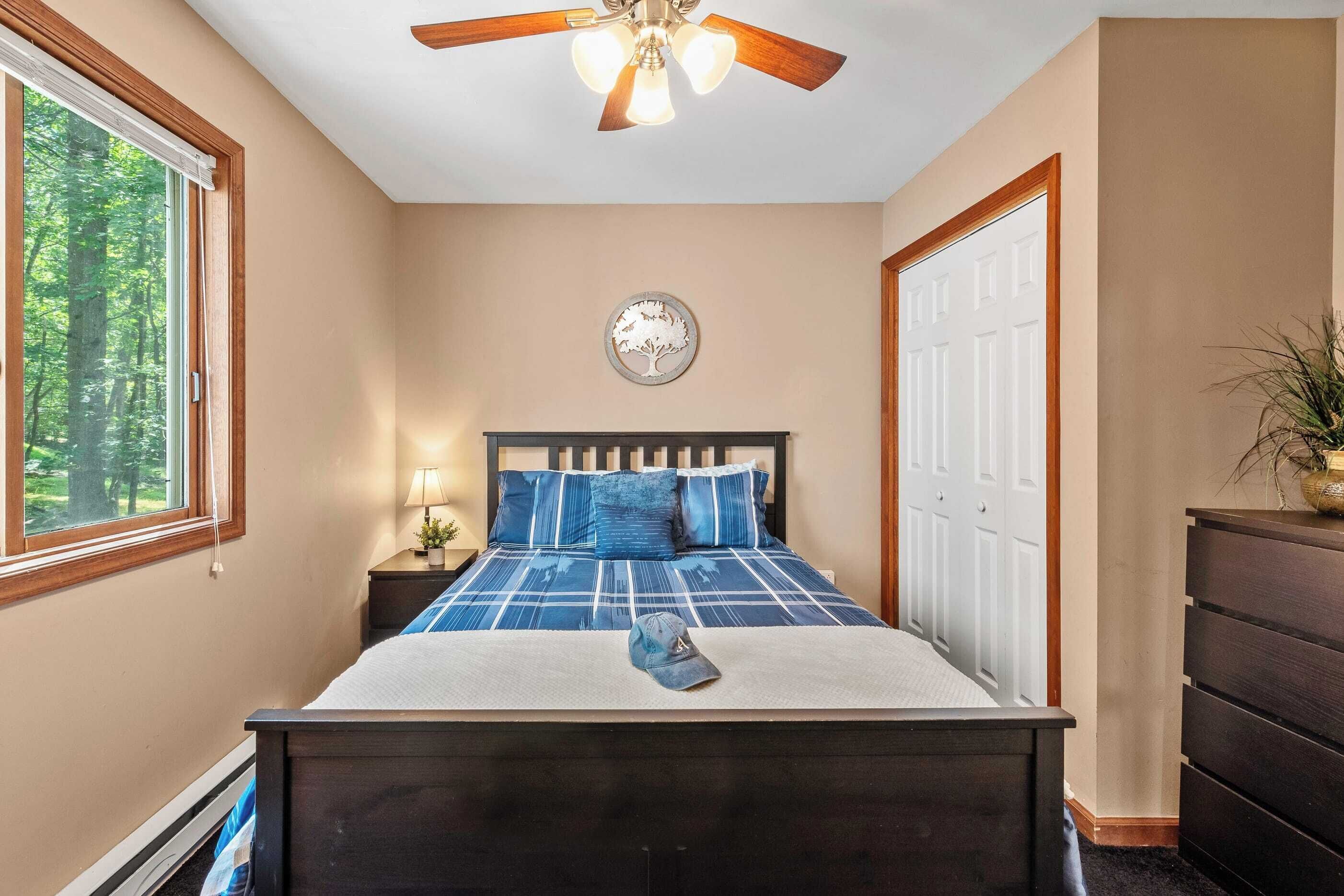 A bedroom with a bed, a ceiling fan, a window, a door, and a wall-mounted clock.