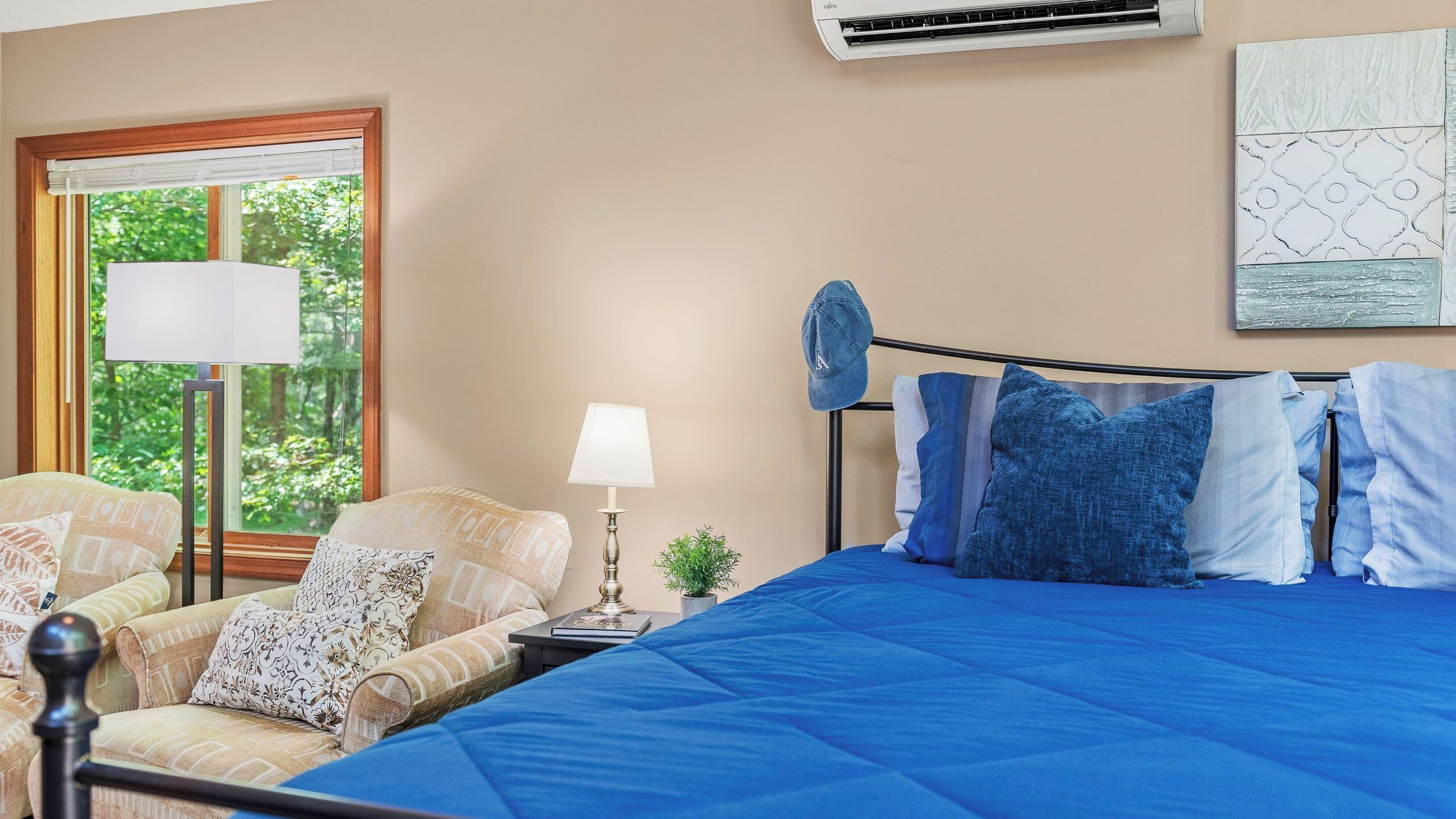 Une chambre à coucher comprenant un lit, une table de chevet, une lampe, une fenêtre donnant sur un espace verdoyant et un climatiseur fixé au mur.