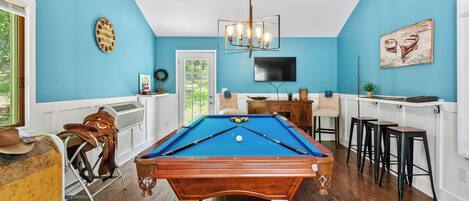 Una mesa de billar con superficie azul de fieltro, una mesa de madera, un reloj, un televisor y una zona de bar con taburetes.