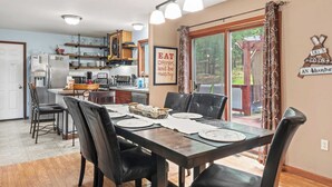 A dining area with a long table set for four, surrounded by black chairs. A kitchen is visible in the background with a refrigerator, stove, and shelves.