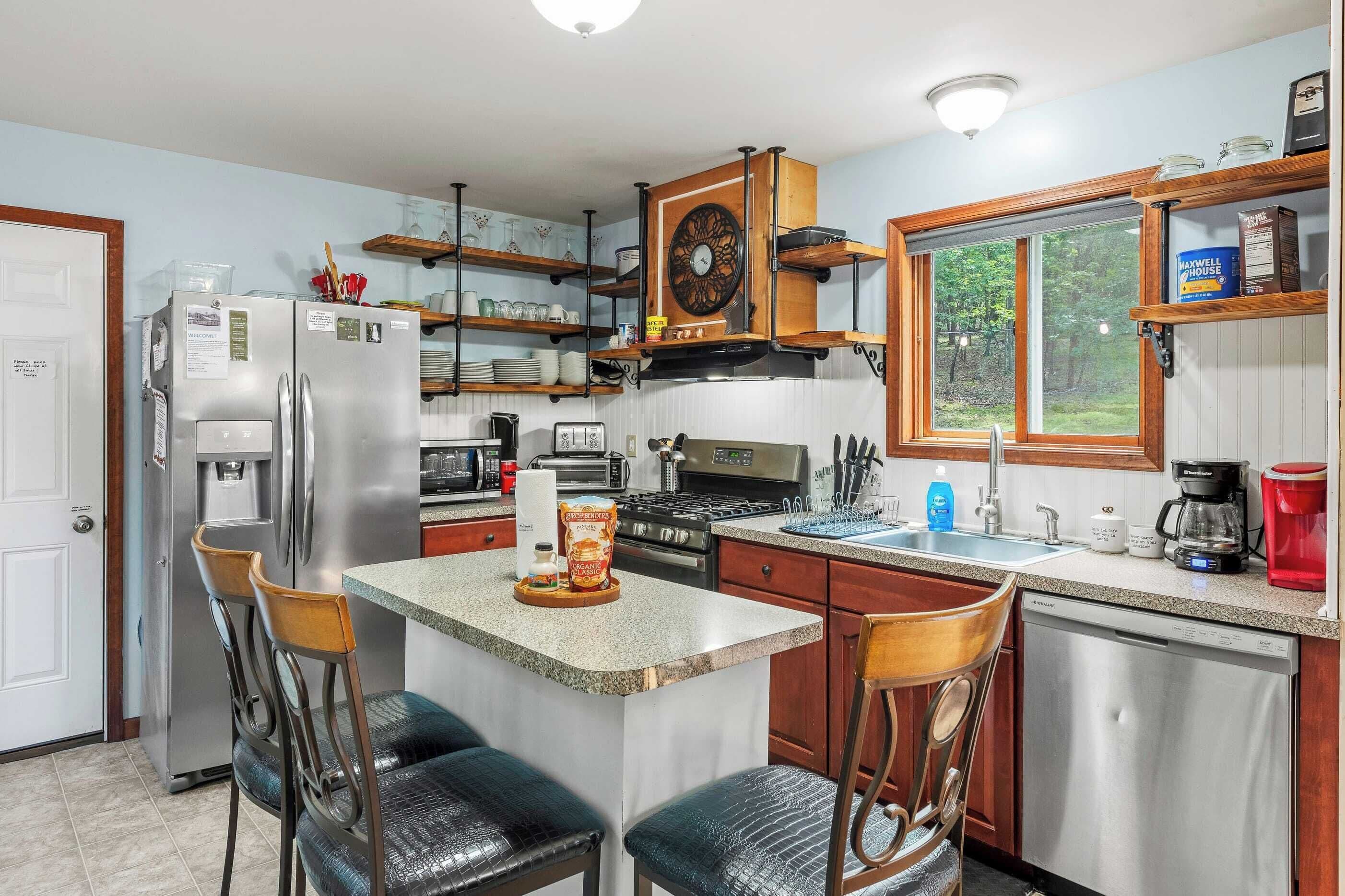 A compact kitchen with a central island, stainless steel appliances, wooden shelves, and a window providing a view of the outdoors.