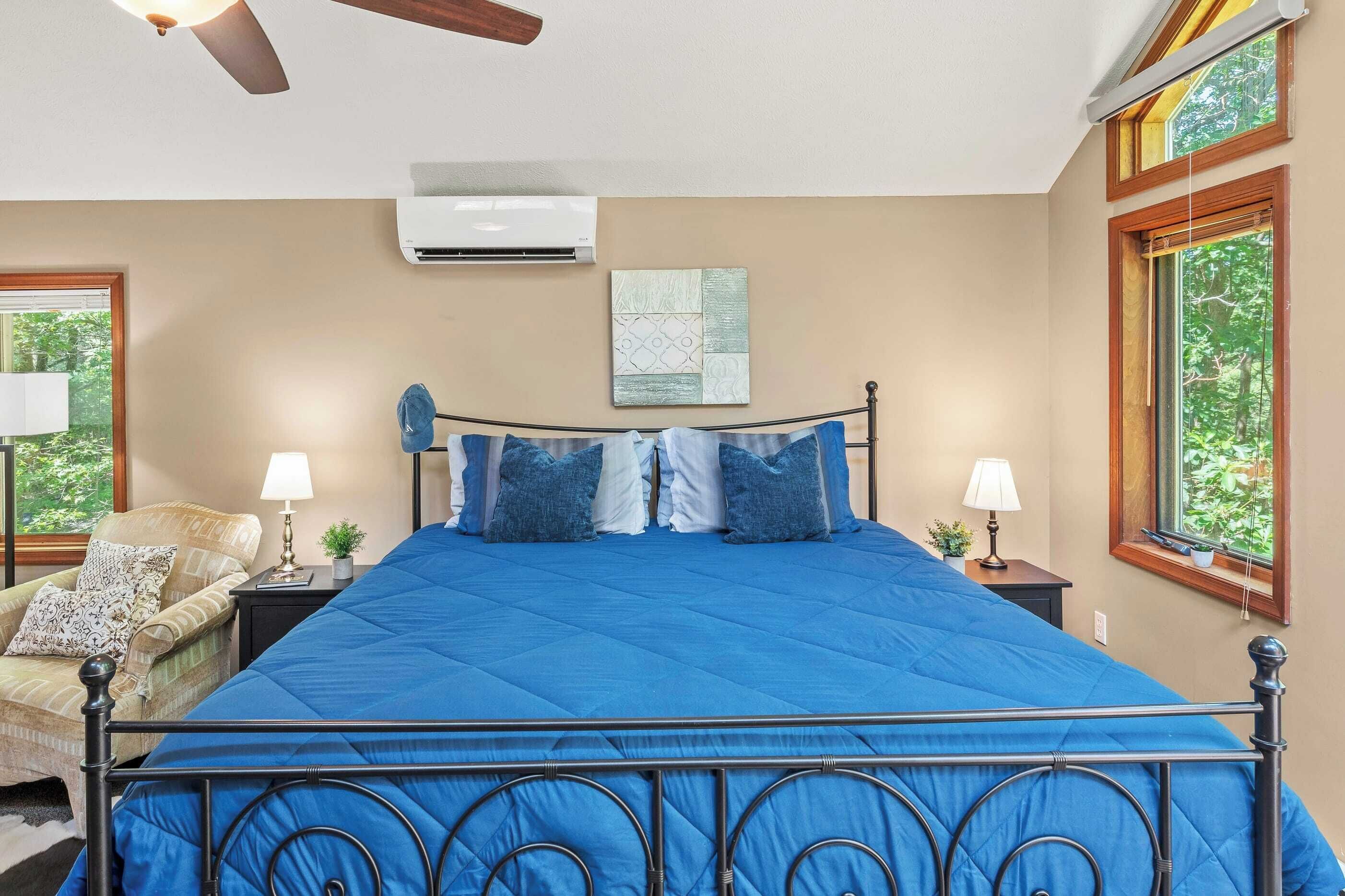 A bedroom with a large bed, two bedside tables with lamps, a chair, a ceiling fan, and a window with a view of greenery.
