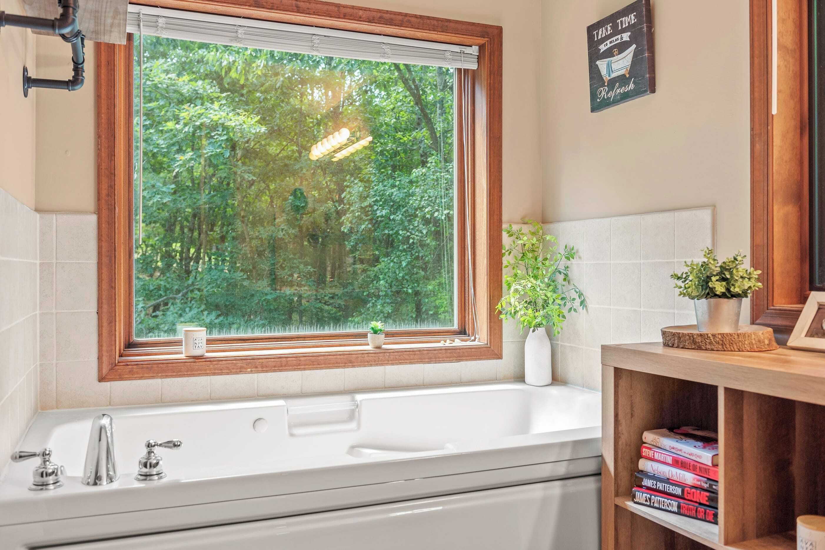 Une salle de bain avec une grande fenêtre, une baignoire, un lavabo avec deux robinets, une plante en pot et une étagère avec des livres.