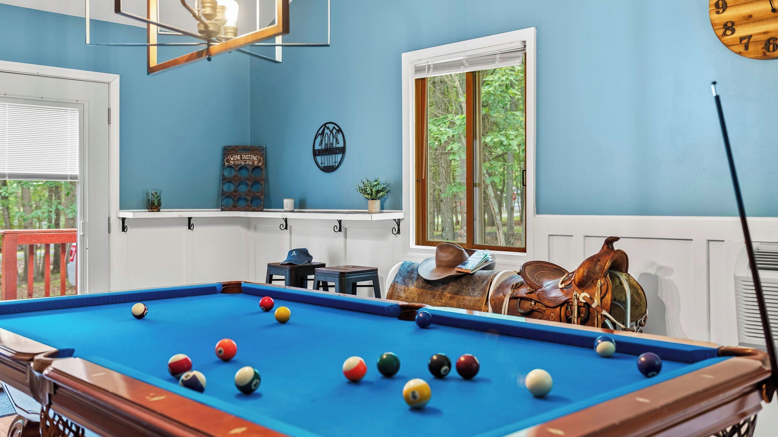 Una mesa de billar con bolas, un reloj, una ventana con vista a árboles y una pared con un letrero que dice 'Cata de Vinos'.