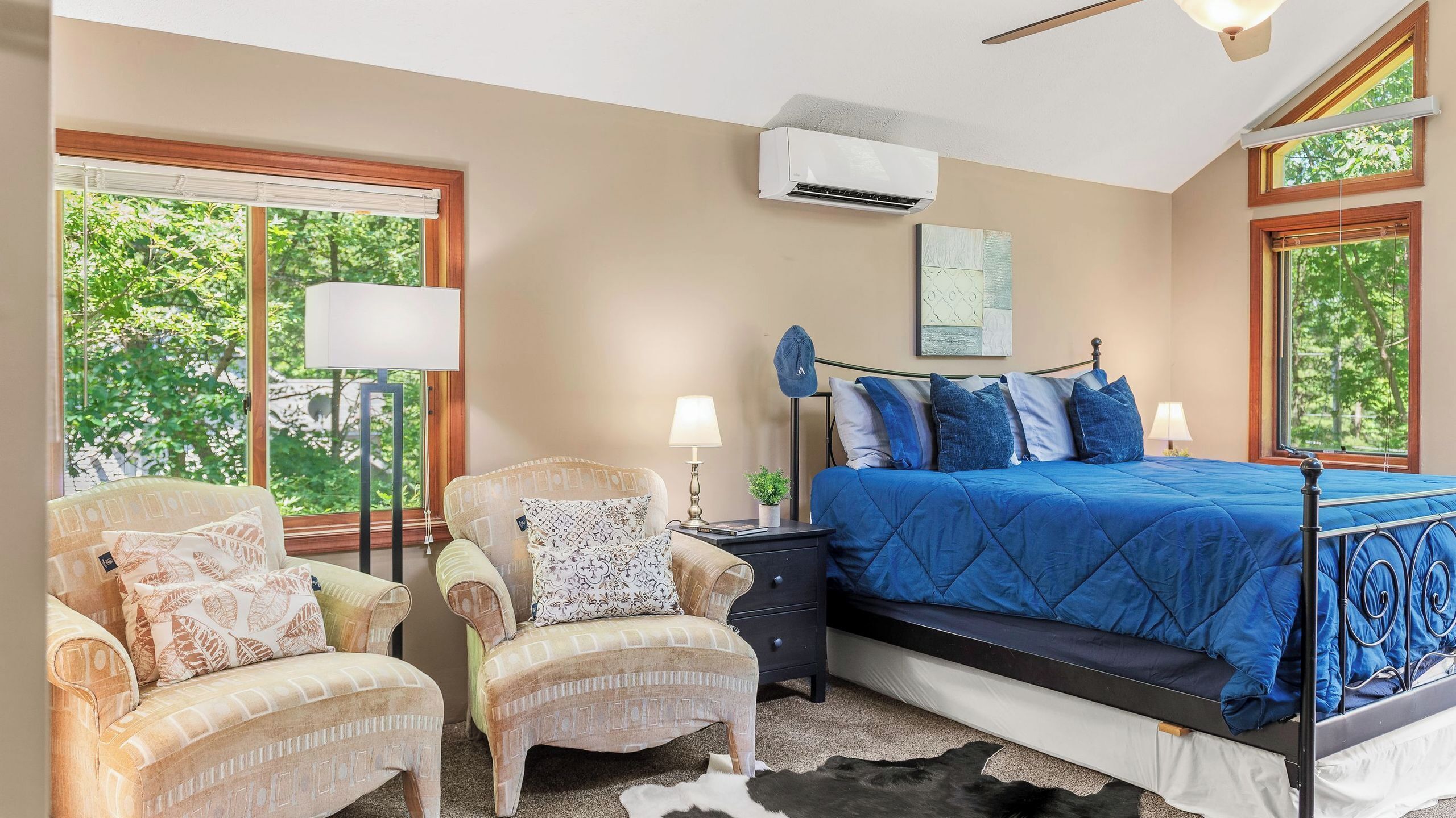 A bedroom with a bed, two armchairs, a nightstand, an air conditioner, and a window with a view of trees.