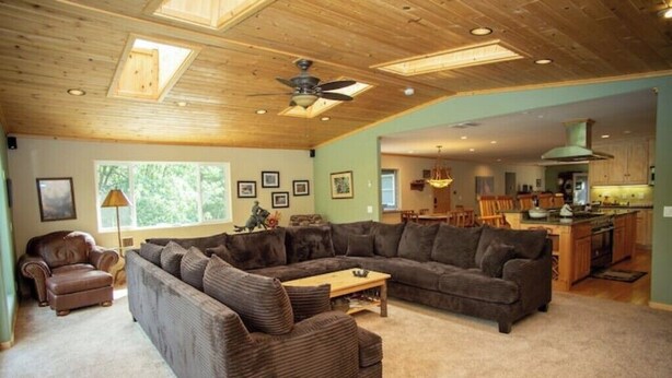 A spacious living room with a large sectional sofa, a small sofa, and a coffee table. There is a dining area with a table and chairs, and a kitchen in the background.