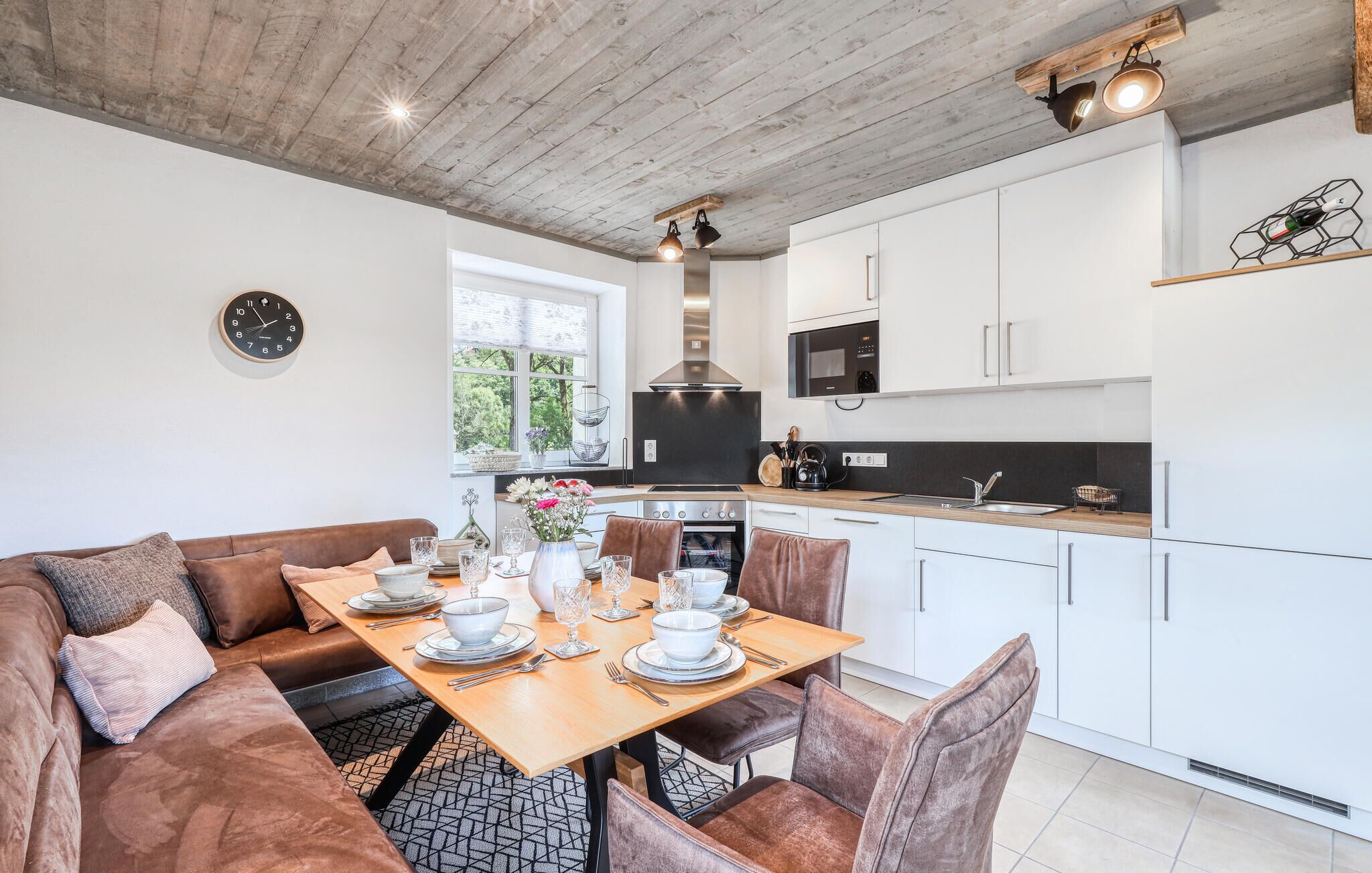 A modern kitchen with white cabinets, a wooden dining table set for four, and a large window providing natural light.