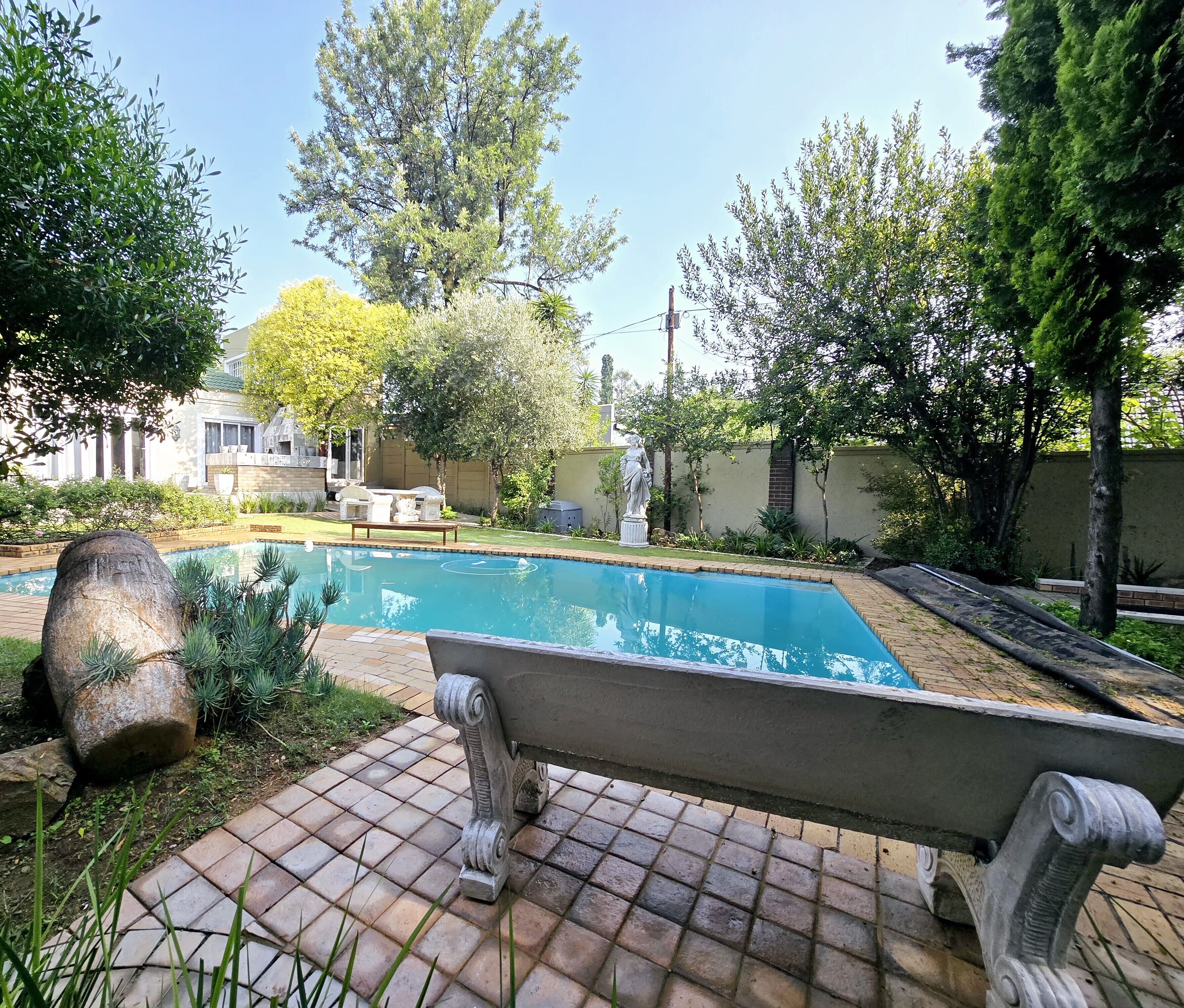 Piscina com um banco de pedra em primeiro plano, cercada por vegetação e um edifício ao fundo.