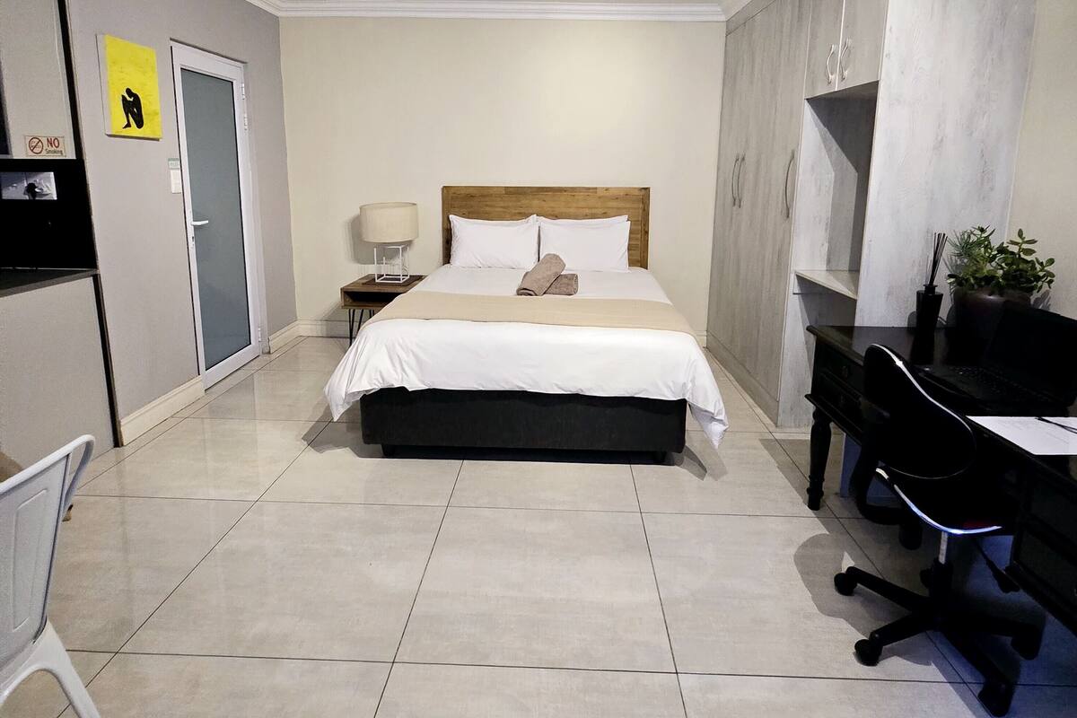 Una habitación de hotel con cama, escritorio, silla, lámpara y una planta.